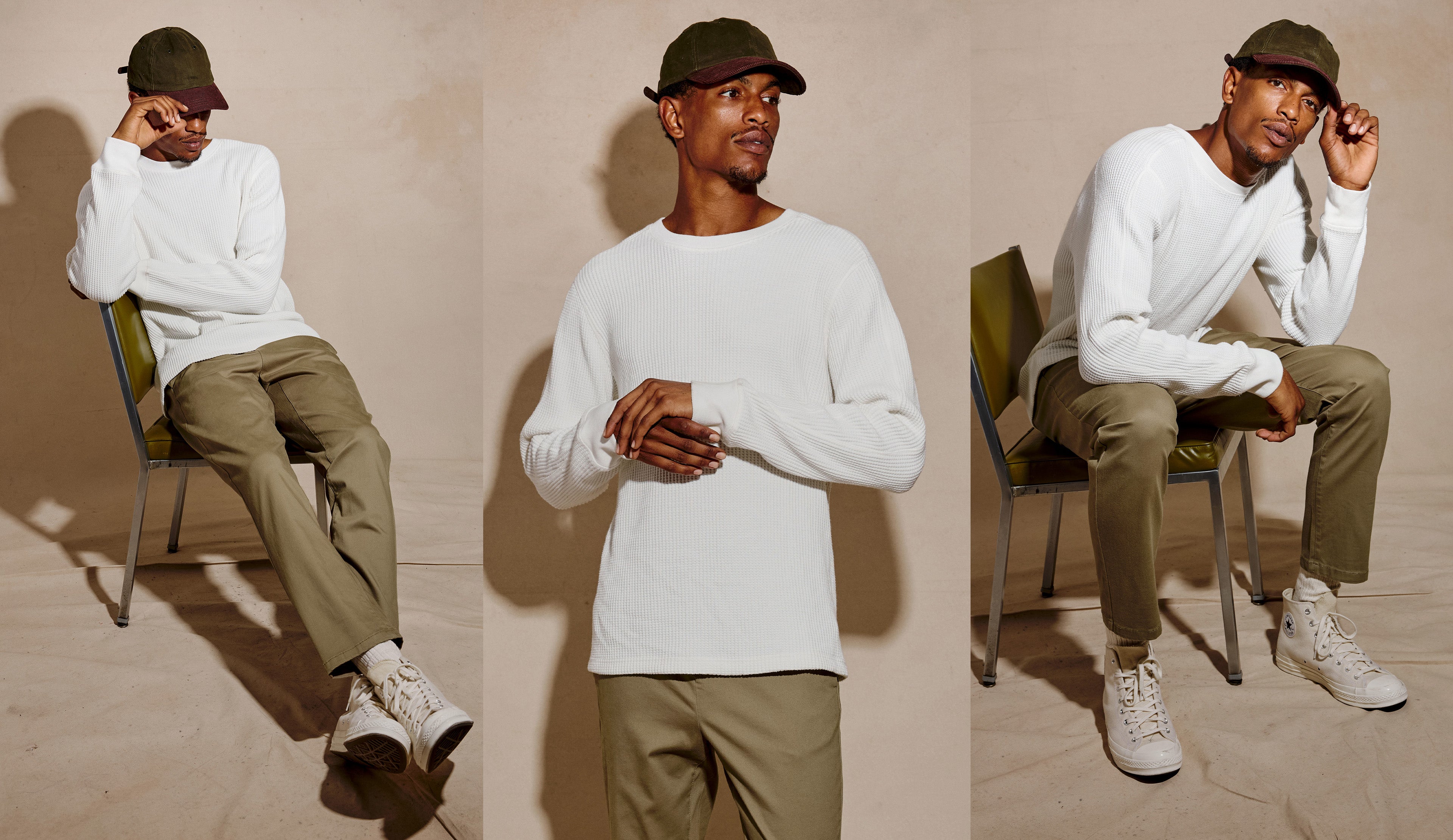 A man sits on a green chair wearing a white long-sleeve crewneck sweatshirt, olive green trousers, white sneakers, and a dark green cap against a beige studio background.
