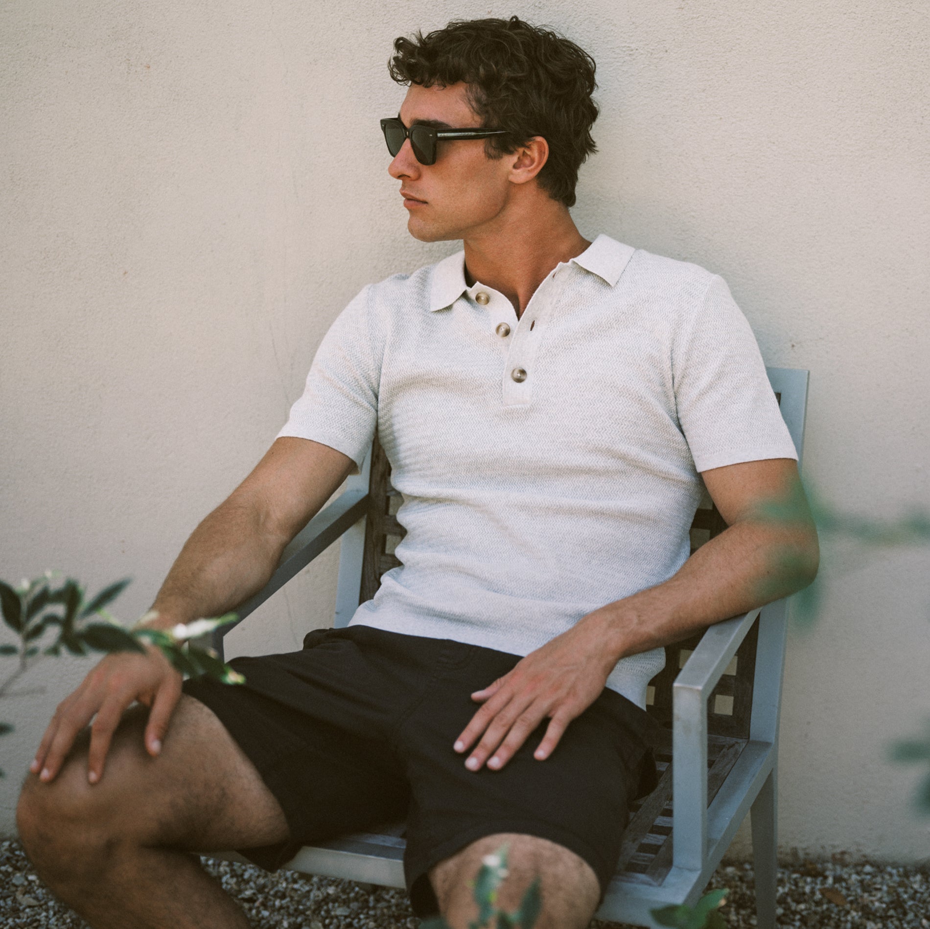 Man wearing sunglasses and a white polo shirt sitting on a chair against a neutral wall.