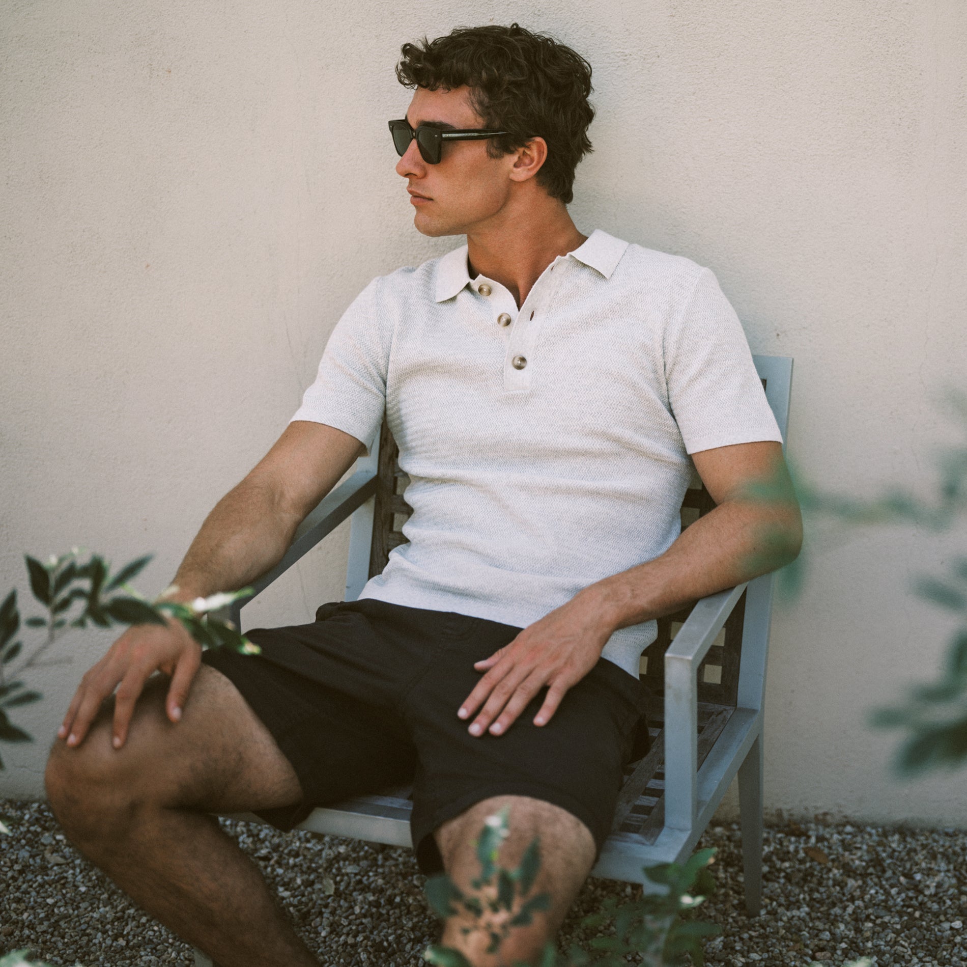 A man wearing a white short-sleeve polo shirt and black shorts sits in a light blue wooden chair against a beige stucco wall, wearing dark sunglasses.