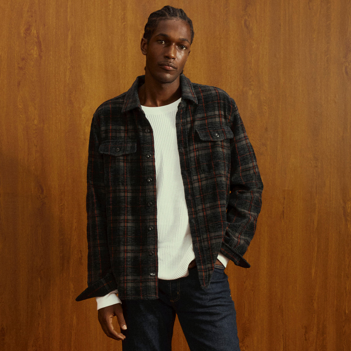 A man wearing a dark plaid button-up shirt over a white ribbed knit sweater and dark blue jeans, standing against a warm wood-paneled background.
