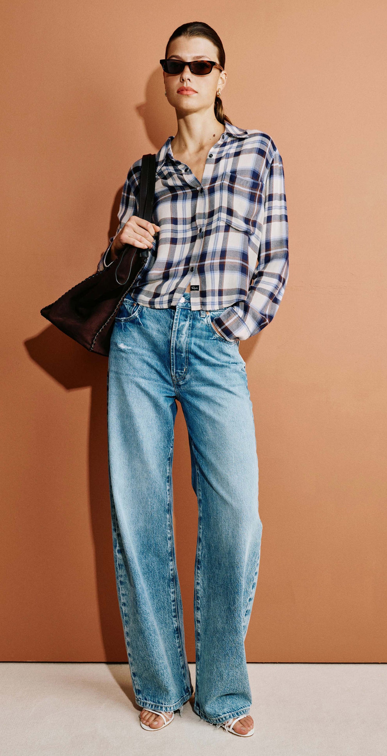 A woman stands against a warm terracotta wall wearing a blue and white plaid button-down shirt, light-wash wide-leg blue jeans, and sunglasses, while carrying a dark shoulder bag.