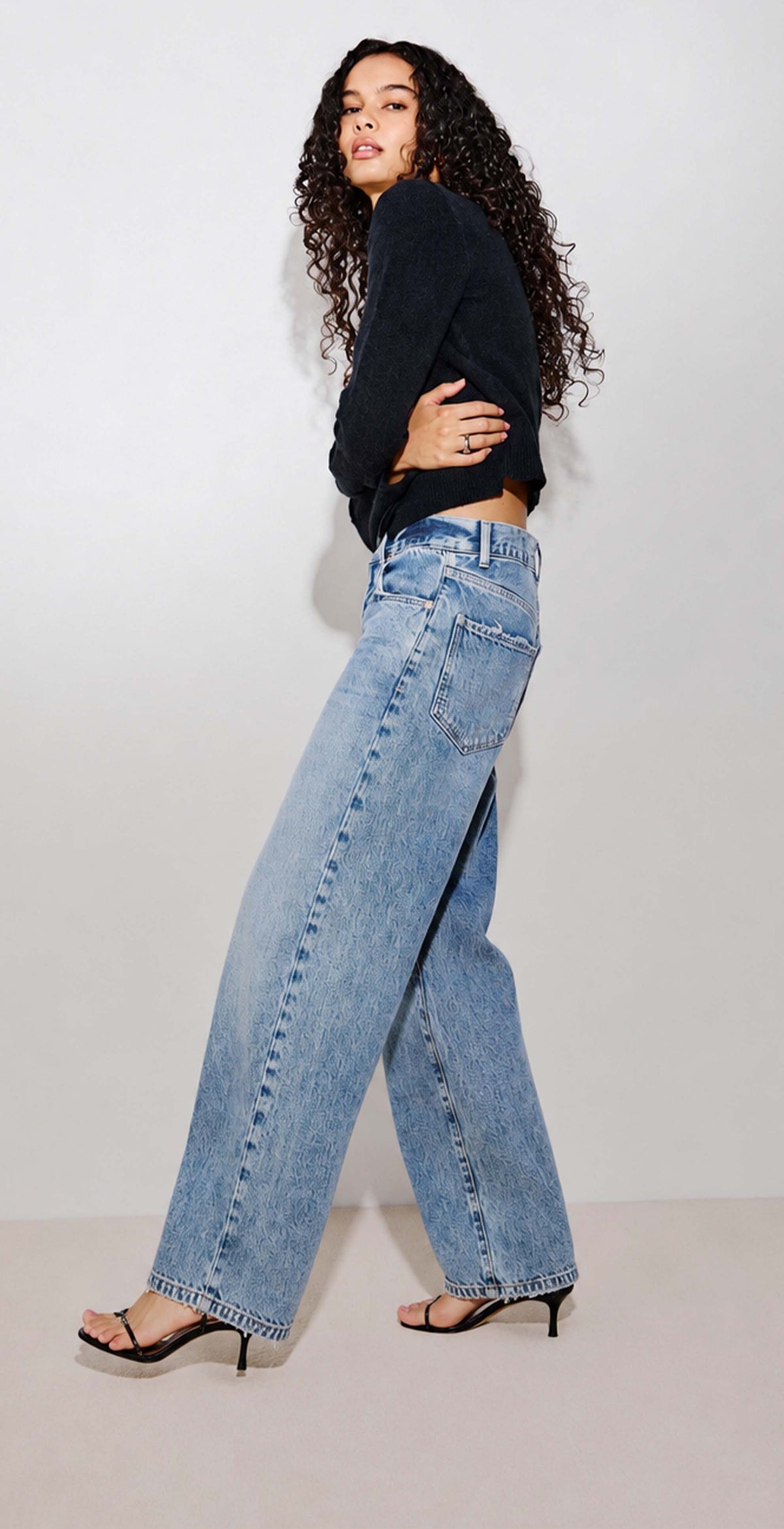 A woman modeling a pair of wide-leg light blue denim jeans with a black cropped long-sleeve top and black strappy heels, standing against a plain off-white studio background.