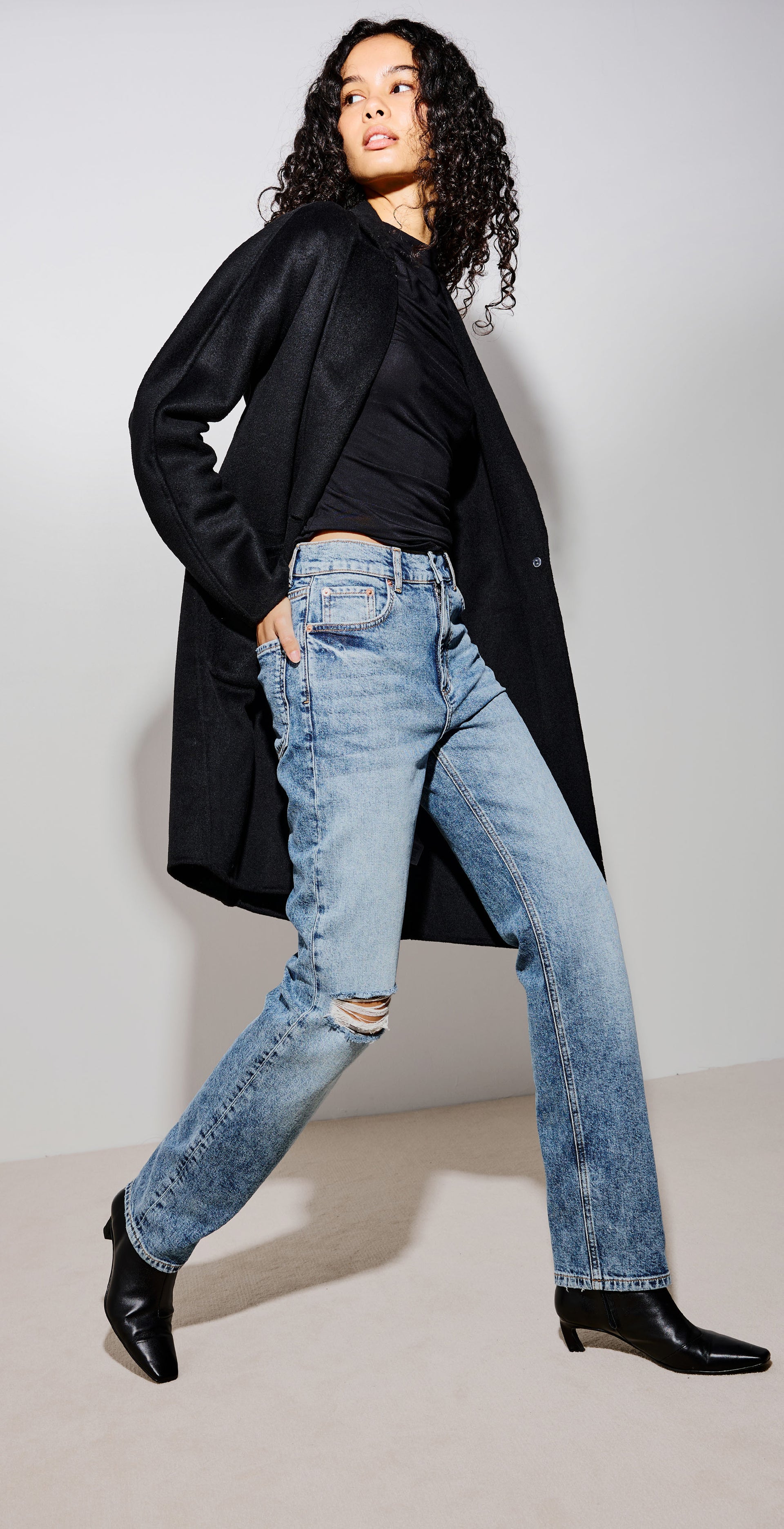 A three-panel studio fashion photo showing a model wearing light-wash distressed blue jeans, a black long-sleeve top, and black ankle boots, posed while sitting on and standing beside a wooden chair against a light gray backdrop.
