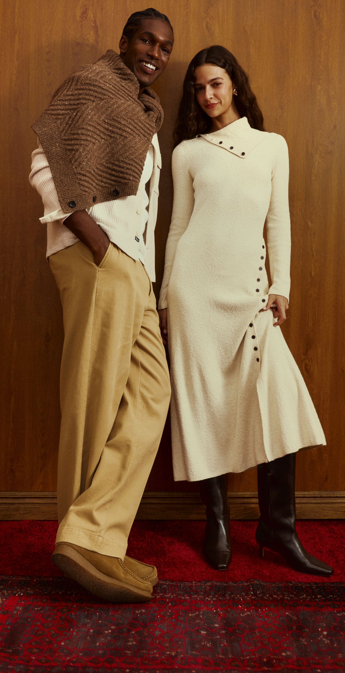 A man and a woman pose together in a warm, rustic indoor setting with a wood-paneled wall and a red patterned rug. The man wears a brown textured knit shawl over a white shirt and tan pleated trousers.