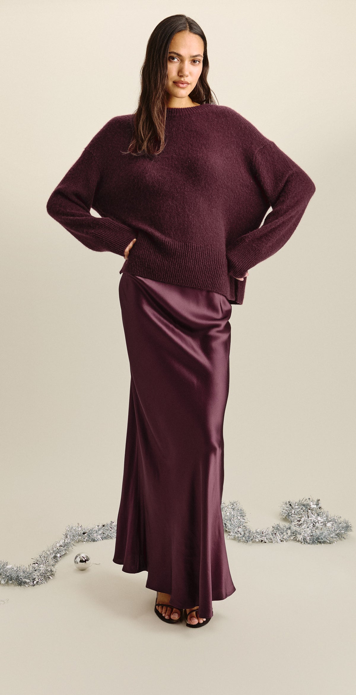A wide triptych in a warm cream studio setting. A model wears an oversized plum knit sweater over a satin plum bias-cut maxi skirt with black strappy heeled sandals. The holiday styling includes silver tinsel, silver ornaments, and a string of vintage multicolored C9 lights draped over her shoulder in the center frame.
