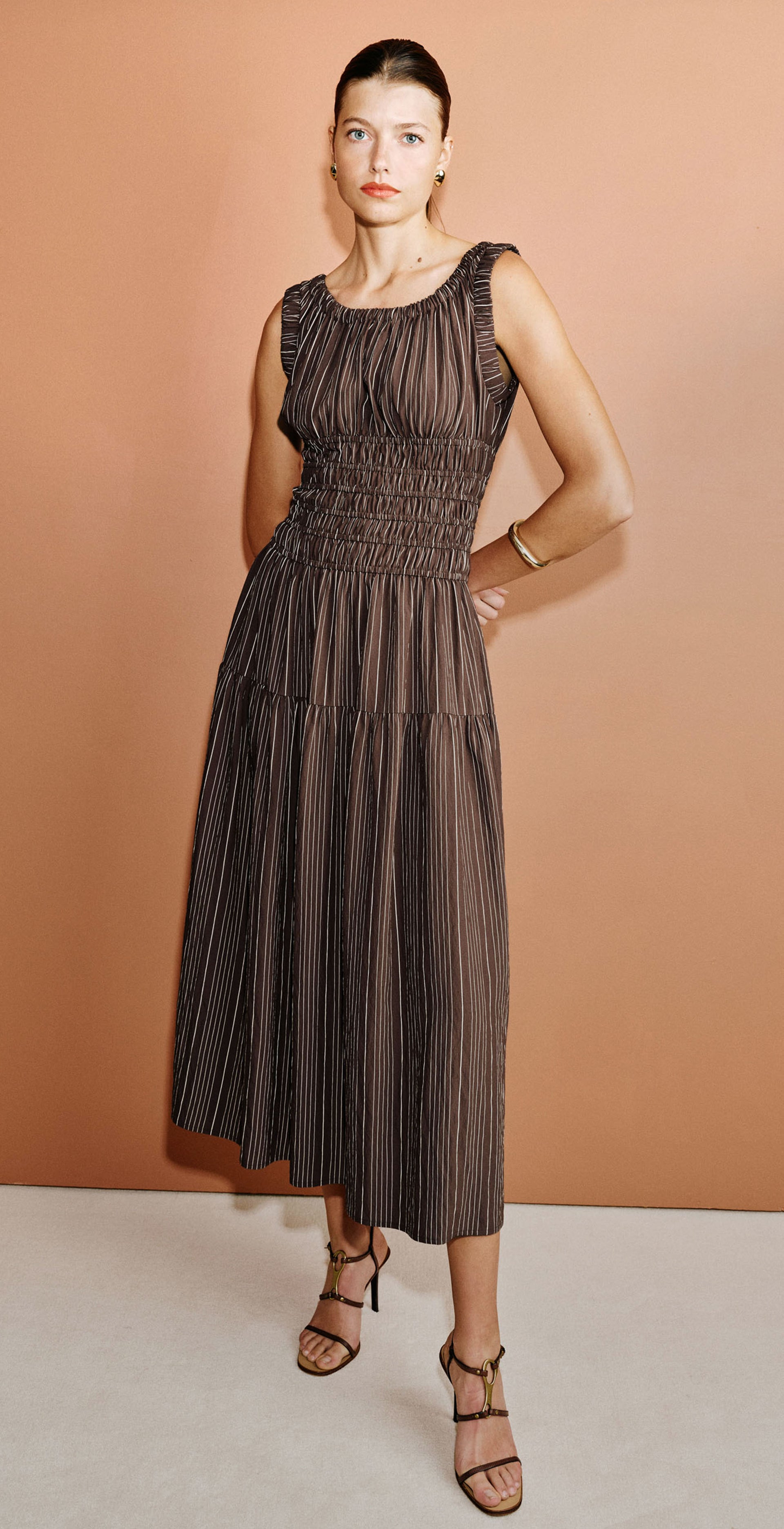 A woman modeling a sleeveless pleated midi dress in a dark brownish-gray color, shown in three poses against a warm peach studio backdrop: seated on a wooden stool, standing with one hand on her hip, and standing with arms crossed.