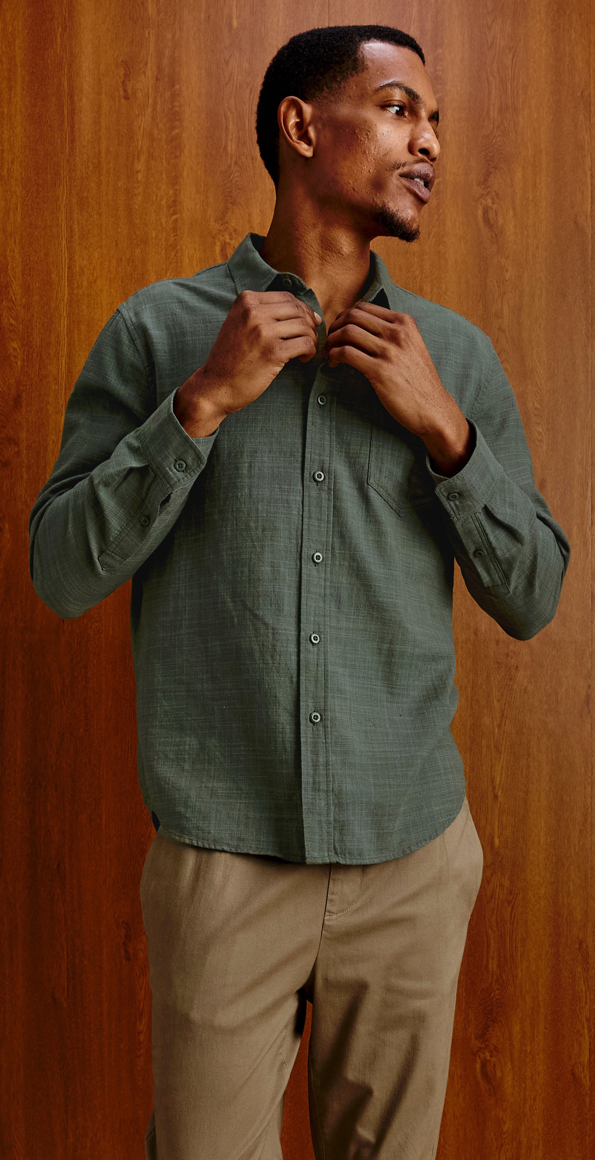 A three-panel lifestyle photo of a man wearing a sage green long-sleeve button-down shirt and khaki pants, shown in different poses against a warm wood-paneled studio background.