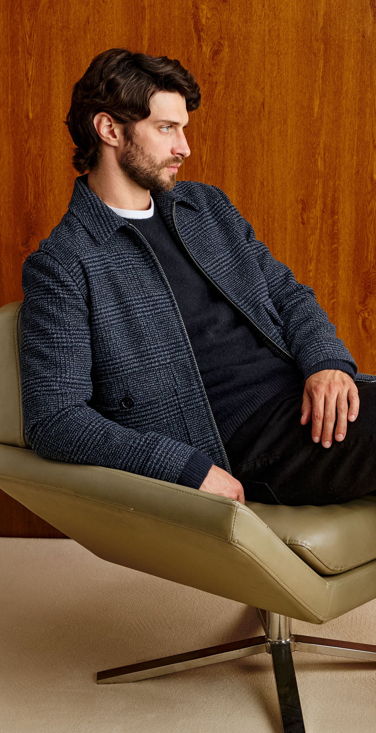A man with dark hair and a beard sits in a beige armchair against a warm wood-paneled wall, wearing a dark blue textured jacket over a black shirt and black trousers.