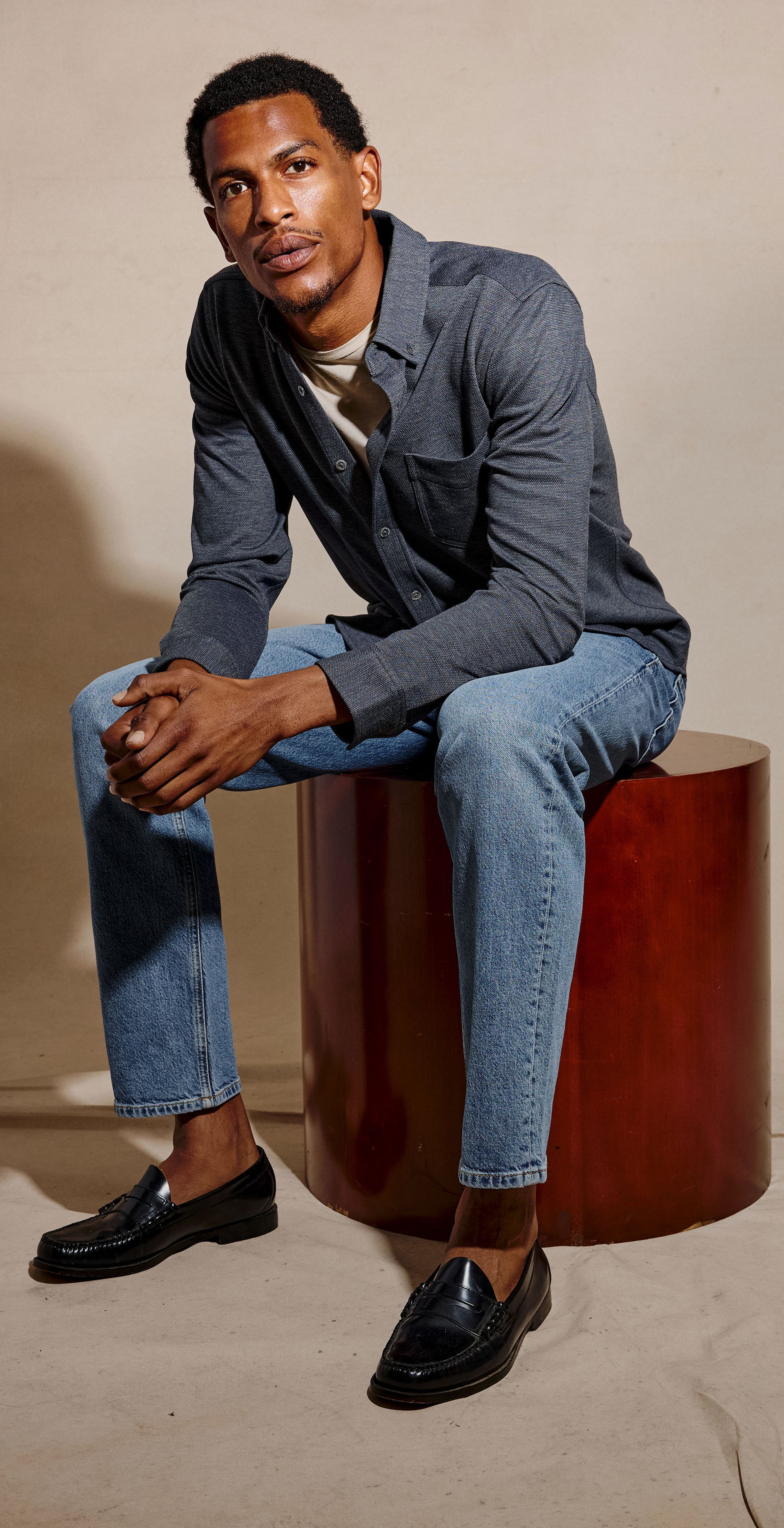 A three-panel studio fashion photo of a man wearing a dark blue-gray long-sleeve button-up shirt with light blue jeans. In the left panel, he stands and adjusts the shirt collar.