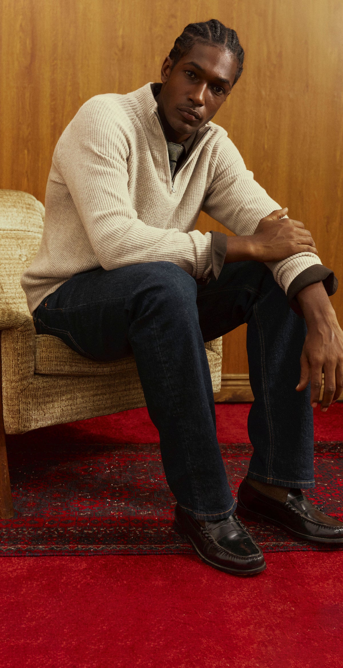 Two shots of a man in a cream waffle-knit quarter-zip sweater over a dark collared shirt and tie, with dark denim; one leaning against a wood-paneled wall, one seated in a vintage armchair on a red rug in leather loafers.