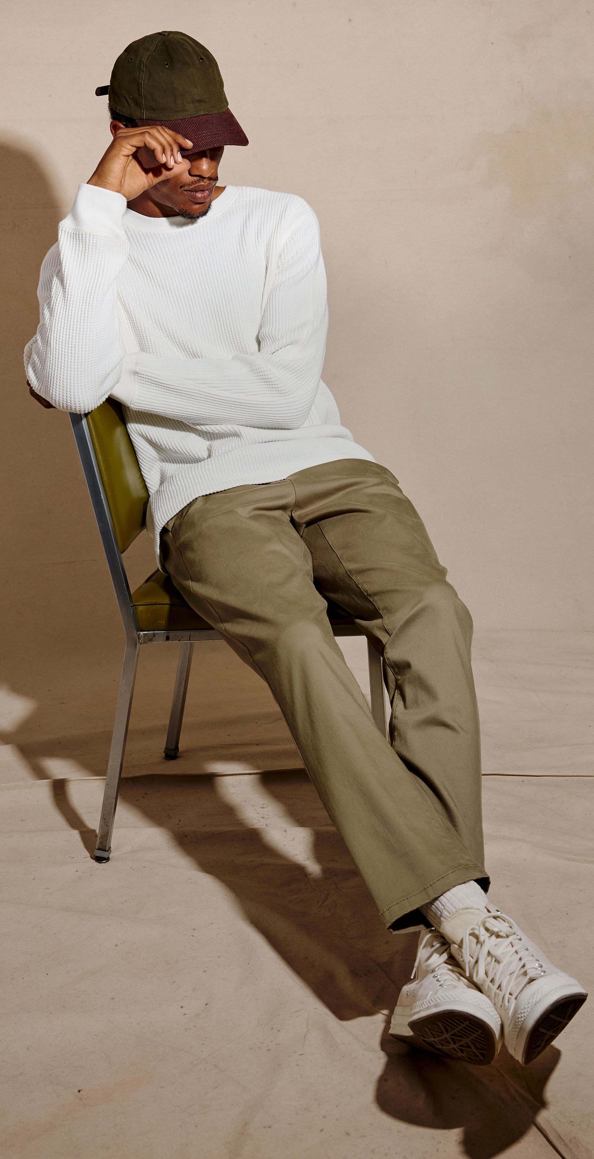 A man sits on a green chair wearing a white long-sleeve crewneck sweatshirt, olive green trousers, white sneakers, and a dark green cap against a beige studio background.