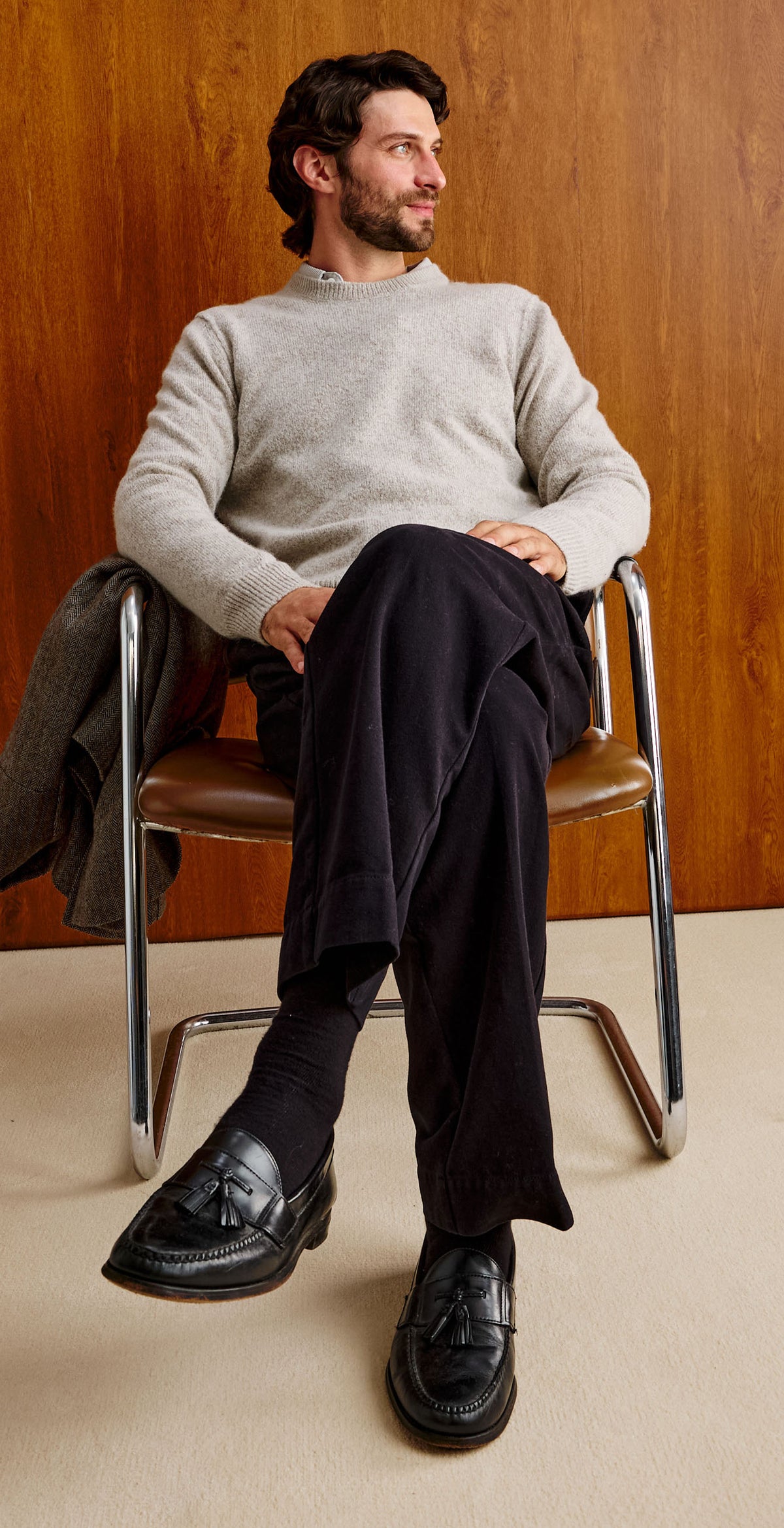 A man with dark hair and a beard sits in a chair against a warm wood-paneled wall, wearing a light gray knit sweater and dark trousers.