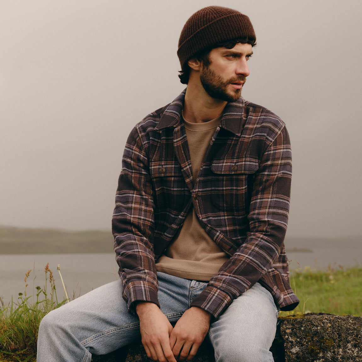 A man wearing a brown knit beanie, a brown and black plaid flannel shirt over a beige t-shirt, and light blue jeans sits on a stone wall outdoors with a misty lake and grassy hills in the background.