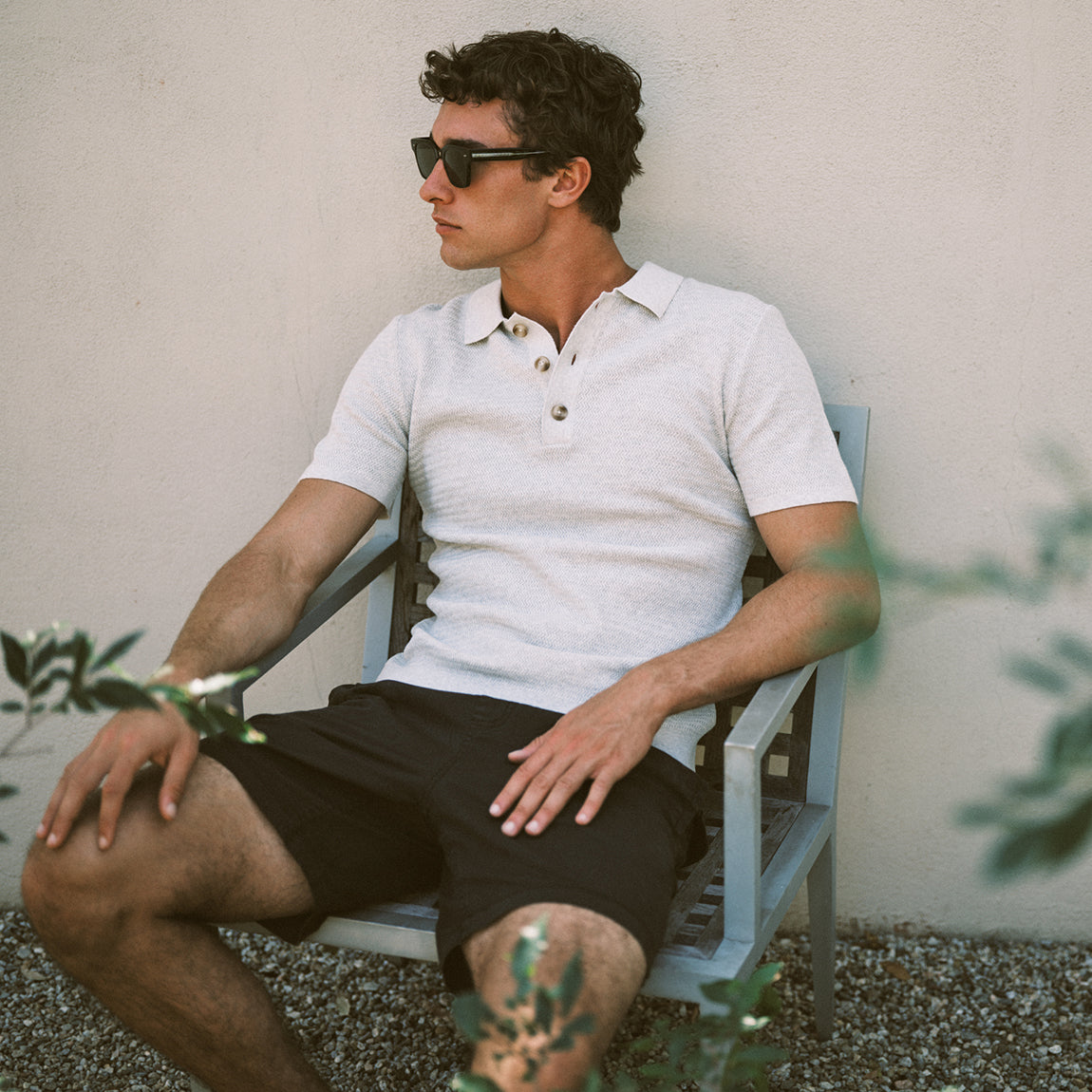 A man wearing a white short-sleeve polo shirt and black shorts sits on a light blue chair against a beige stucco wall, wearing dark sunglasses.