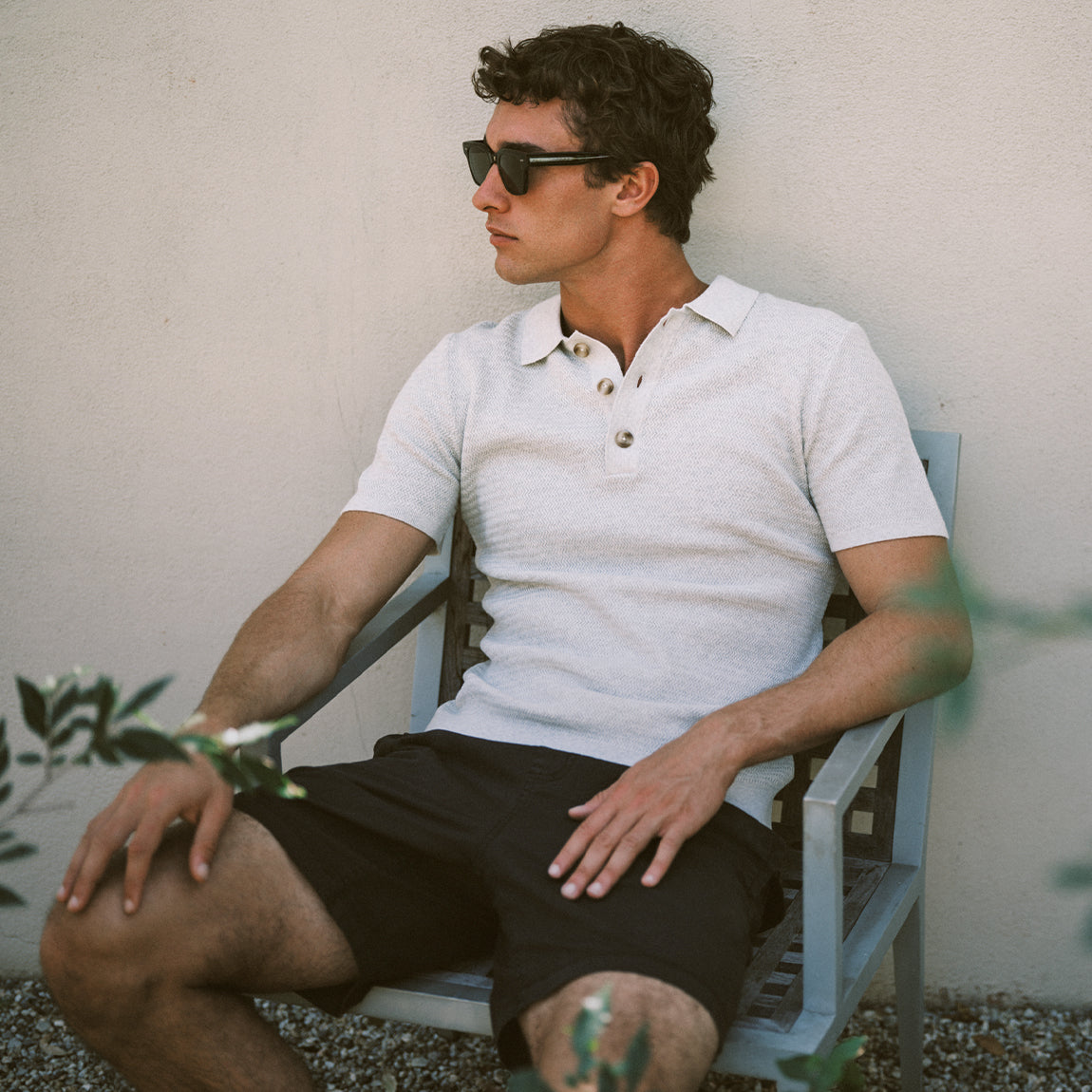 Man wearing sunglasses and a white polo shirt sitting on a chair against a neutral wall.