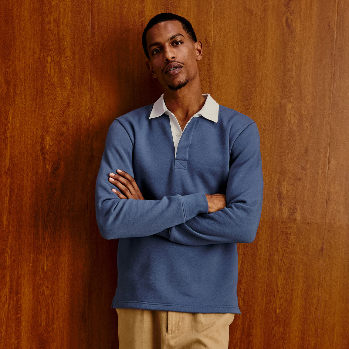 A man wearing a slate blue long-sleeve rugby-style polo shirt with a white collar, standing with arms crossed against a warm wood-paneled background.