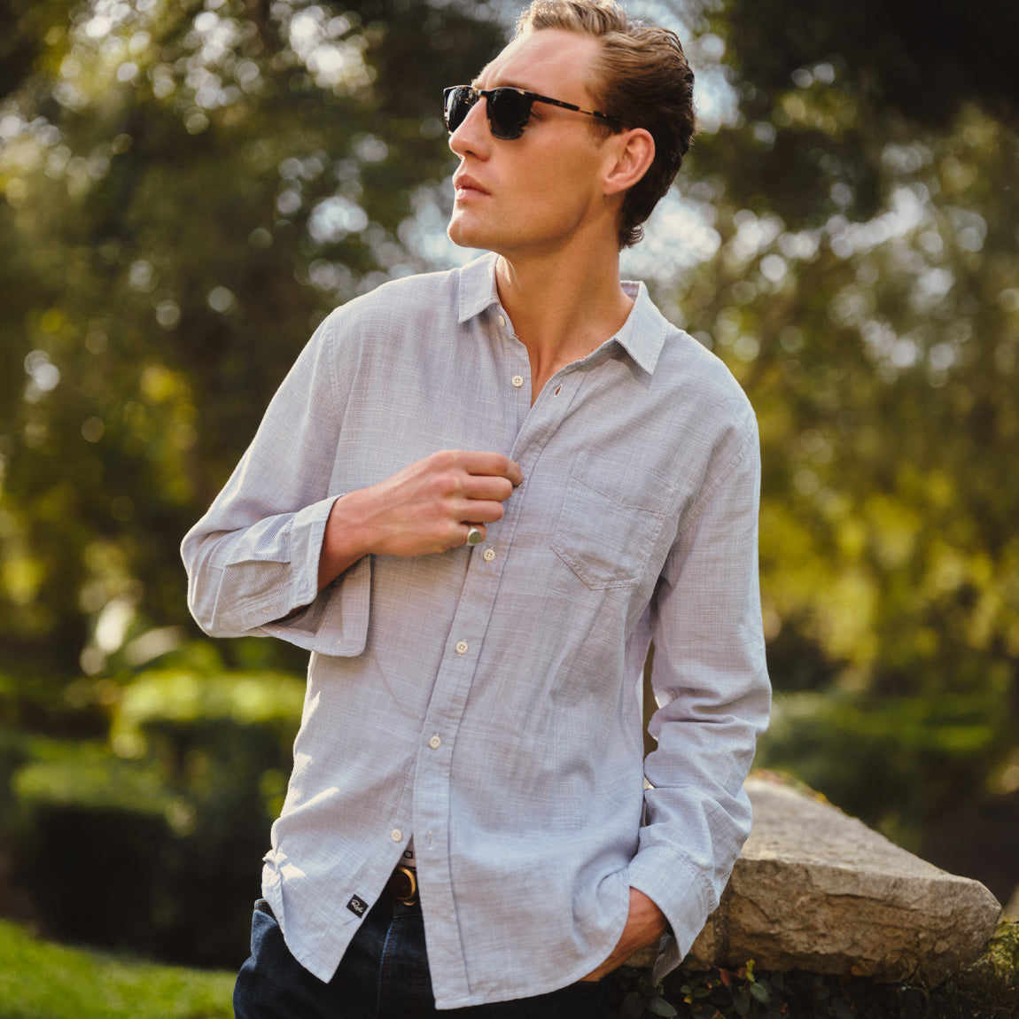Man wearing sunglasses and a light gray shirt outdoors with greenery in the background
