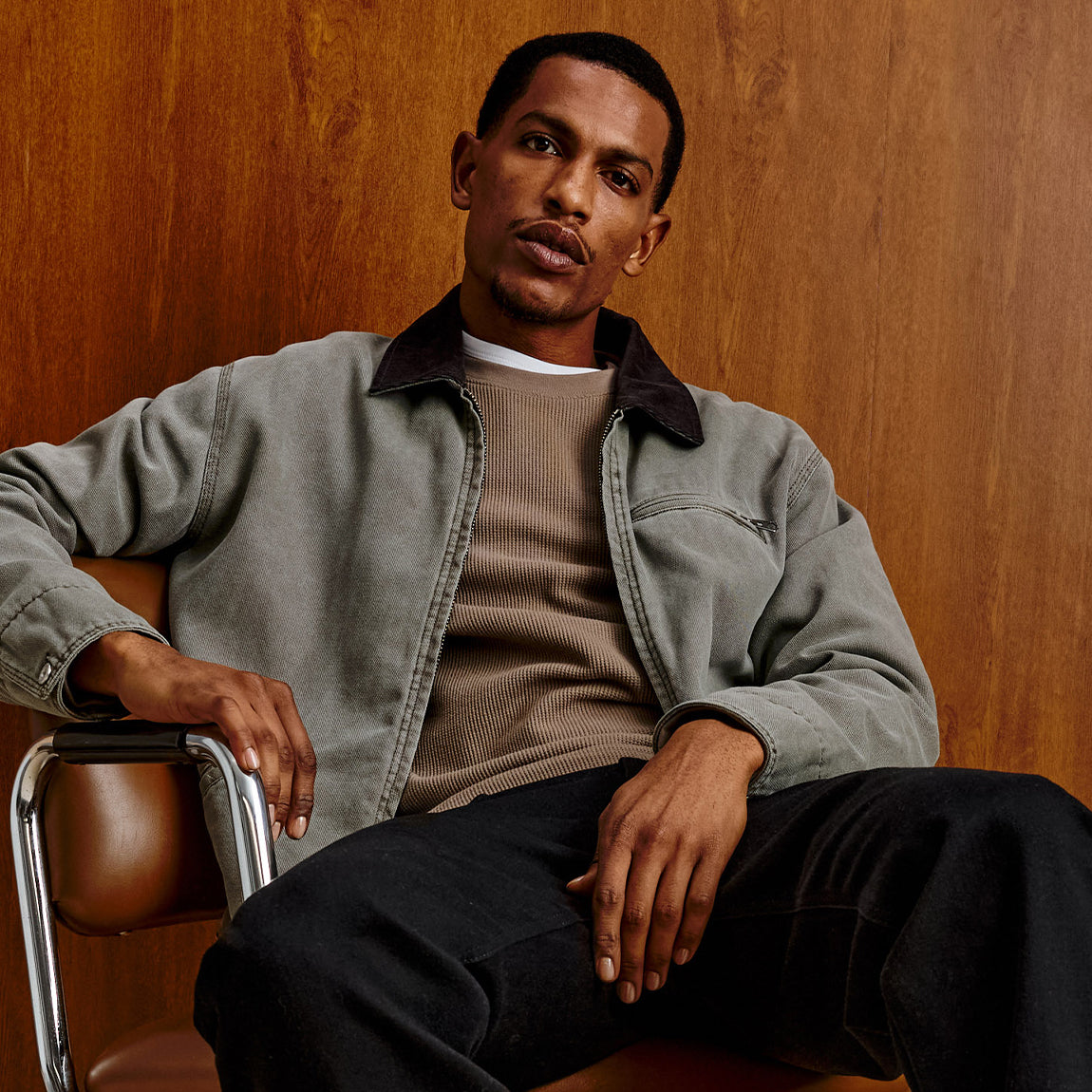 A man wearing a muted gray-green overshirt over a taupe knit sweater and black pants, seated on a brown leather chair against a warm wood-paneled wall