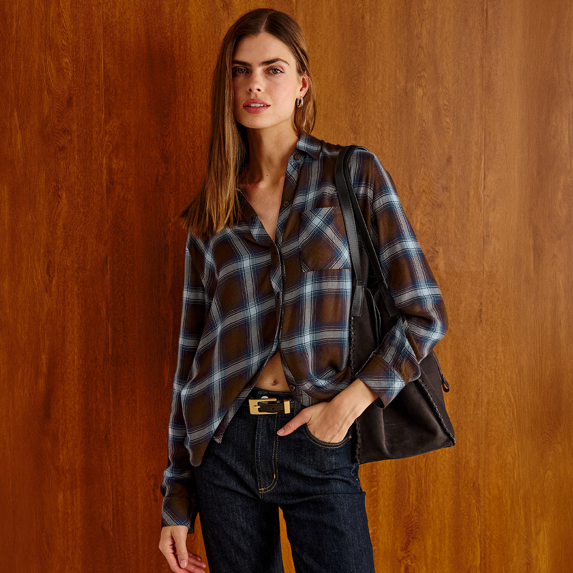 A woman stands against a warm wood-paneled wall wearing a brown and blue plaid button-up shirt, dark blue jeans, and a black shoulder bag.