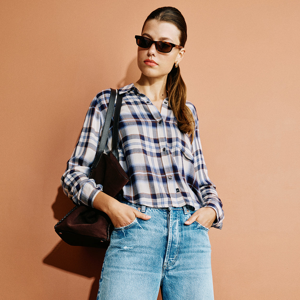 A woman wearing a blue, white, and navy plaid button-down shirt, light-wash blue jeans, and dark sunglasses, carrying a dark brown shoulder bag against a solid terracotta background.