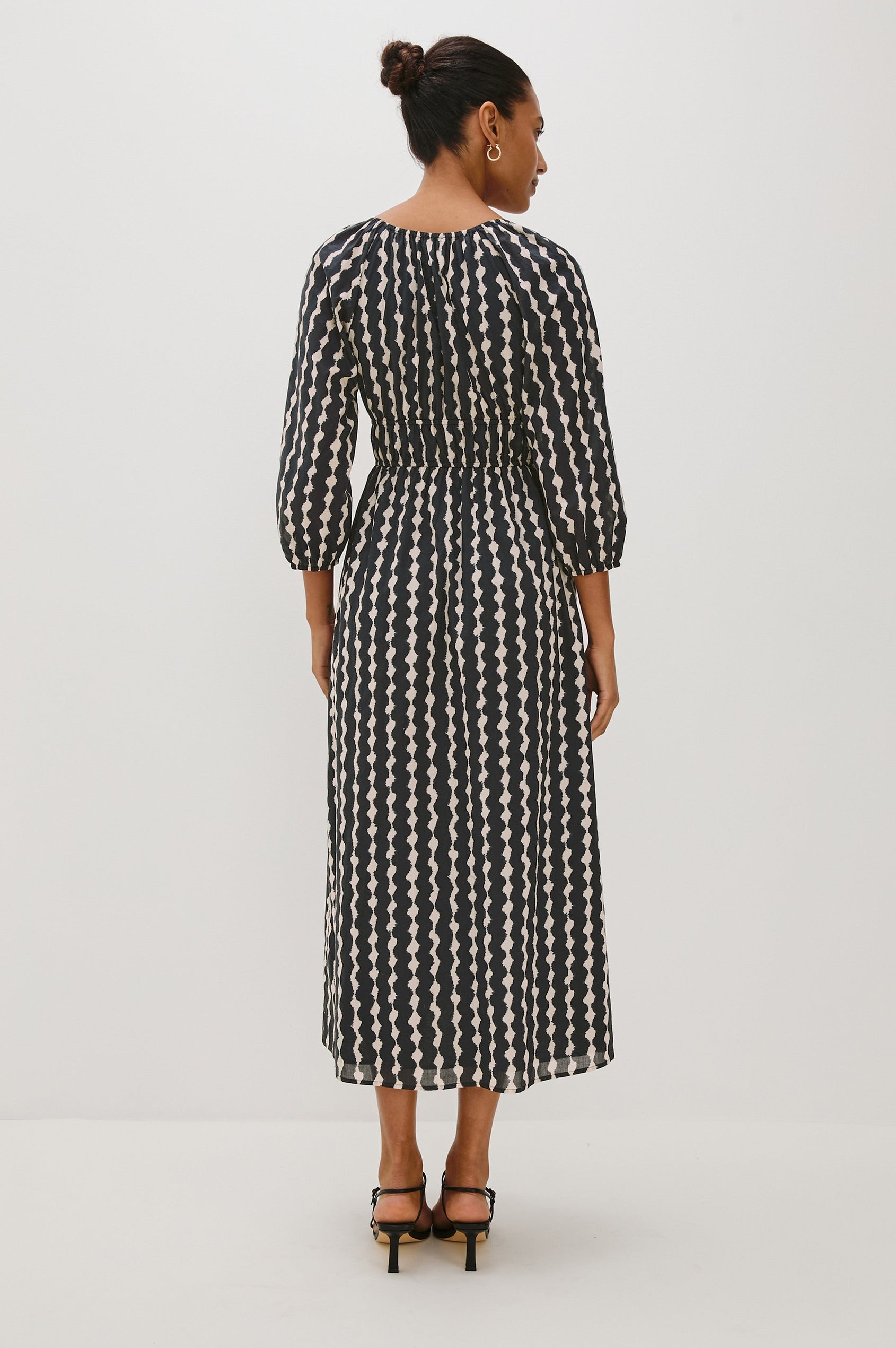 Paio midi dress with a fitted bodice, three quarter sleeves with a slight puff effect, flowing skirt, and eyelet closures at the chest in Painted Noir colorway - back full body view on model