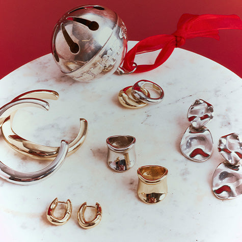 A styled flat-lay of multiple sculptural jewelry pieces in polished silver and gold finishes, including a large silver cuff, a silver bell with a red ribbon, several rings, small cylindrical earrings, and irregular dangling earrings, arranged on a white marble surface against a red background.