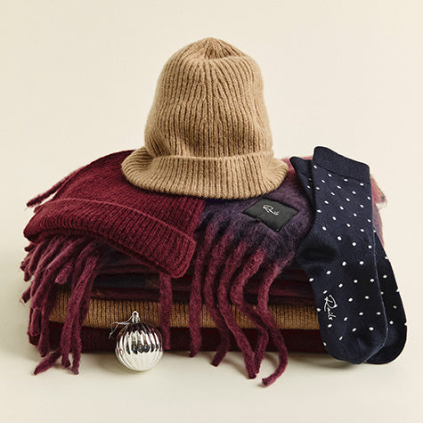 Camel ribbed beanie resting on a stack of fringed scarves in burgundy and mustard, beside a navy polka-dot Rails sock and a silver ornament, on a cream background.