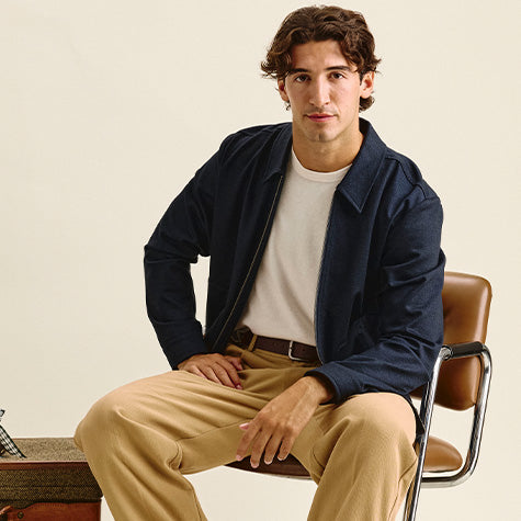A man sits on a brown leather chair wearing a navy blue zip-up jacket over a cream t-shirt and beige trousers, against a plain beige background.