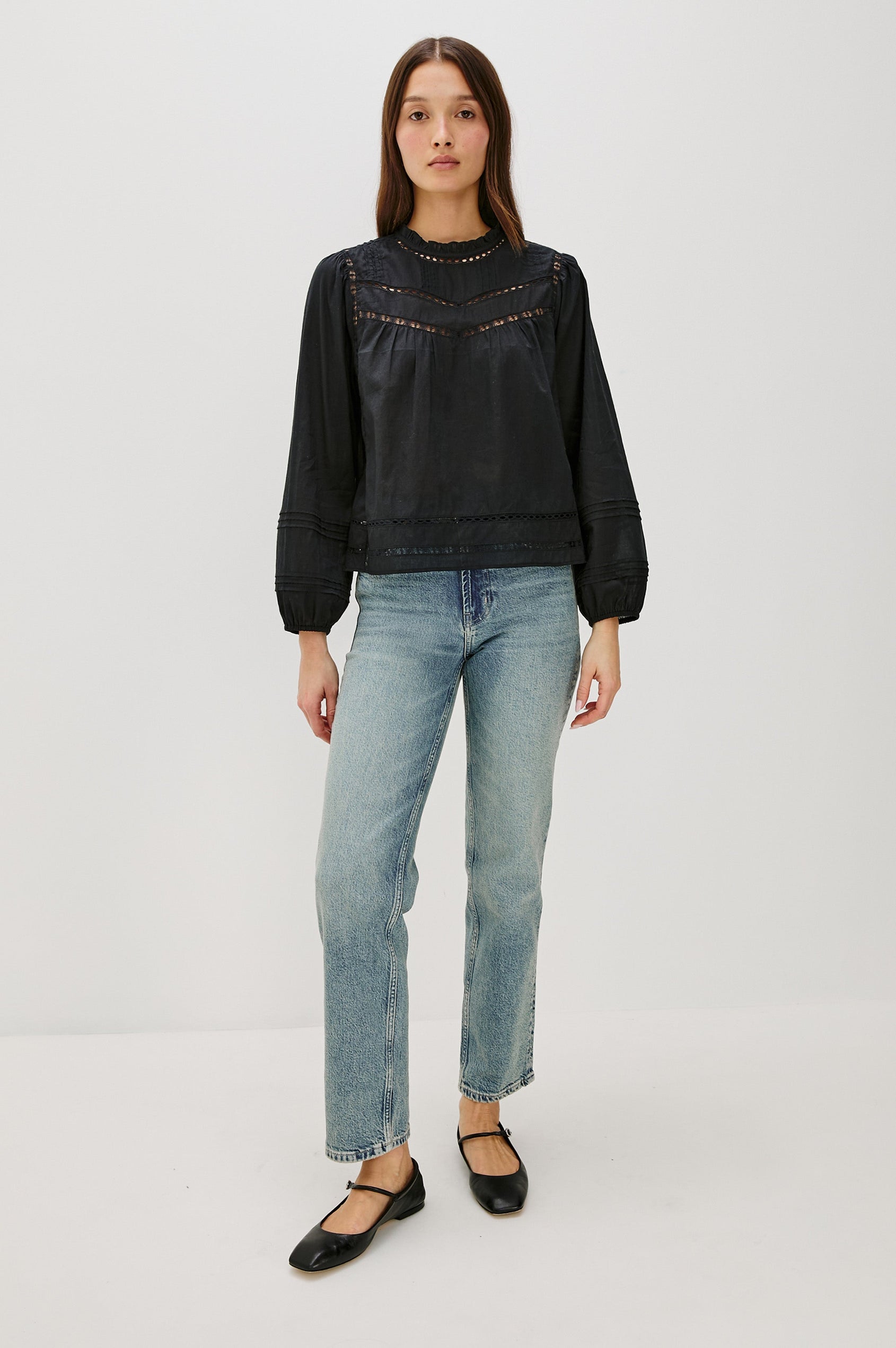 Front full body view of female model wearing our Reyna Top in black. This is a long sleeve crewneck baby doll blouse, with ruffles at the collar. Styled with light wash denim.