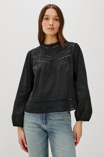 Front view of female model wearing our Reyna Top in black. This is a long sleeve crewneck baby doll blouse, with ruffles at the collar. Styled with light wash denim.