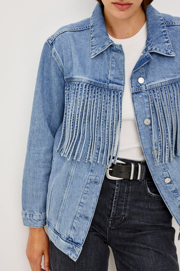 The Rodeo is button front collared denim jacket with side pockets and fringe along front yoke in Indigo Cowboy colorway, a medium blue wash. Front detail view featured on model.
