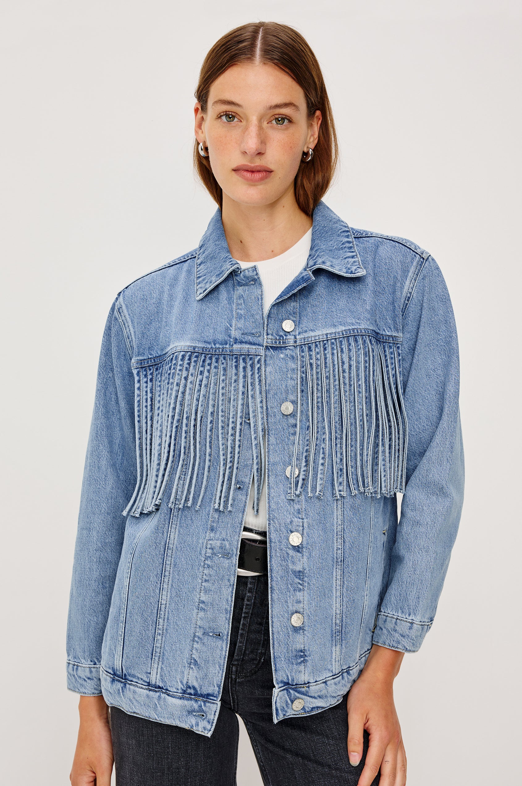 The Rodeo is button front collared denim jacket with side pockets and fringe along front yoke in Indigo Cowboy colorway, a medium blue wash. Front view featured on model.