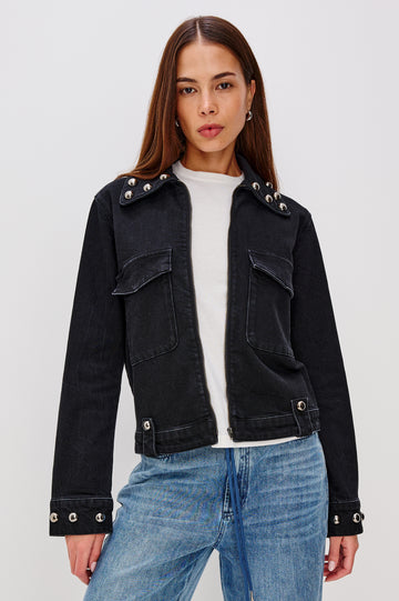 Roxbury is a boxy fit denim jacket featuring nail-back studs at the oversized collar, cuffs, and hem. It has oversized chest pockets with hidden snap closures, hem tabs, and a sleek front zipper closure in Black Stone Studded colorway. Front view featured on model.