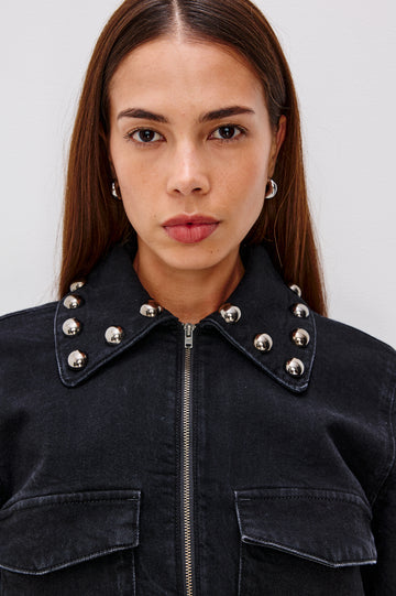Roxbury is a boxy fit denim jacket featuring nail-back studs at the oversized collar, cuffs, and hem. It has oversized chest pockets with hidden snap closures, hem tabs, and a sleek front zipper closure in Black Stone Studded colorway. Front detail view featured on model.