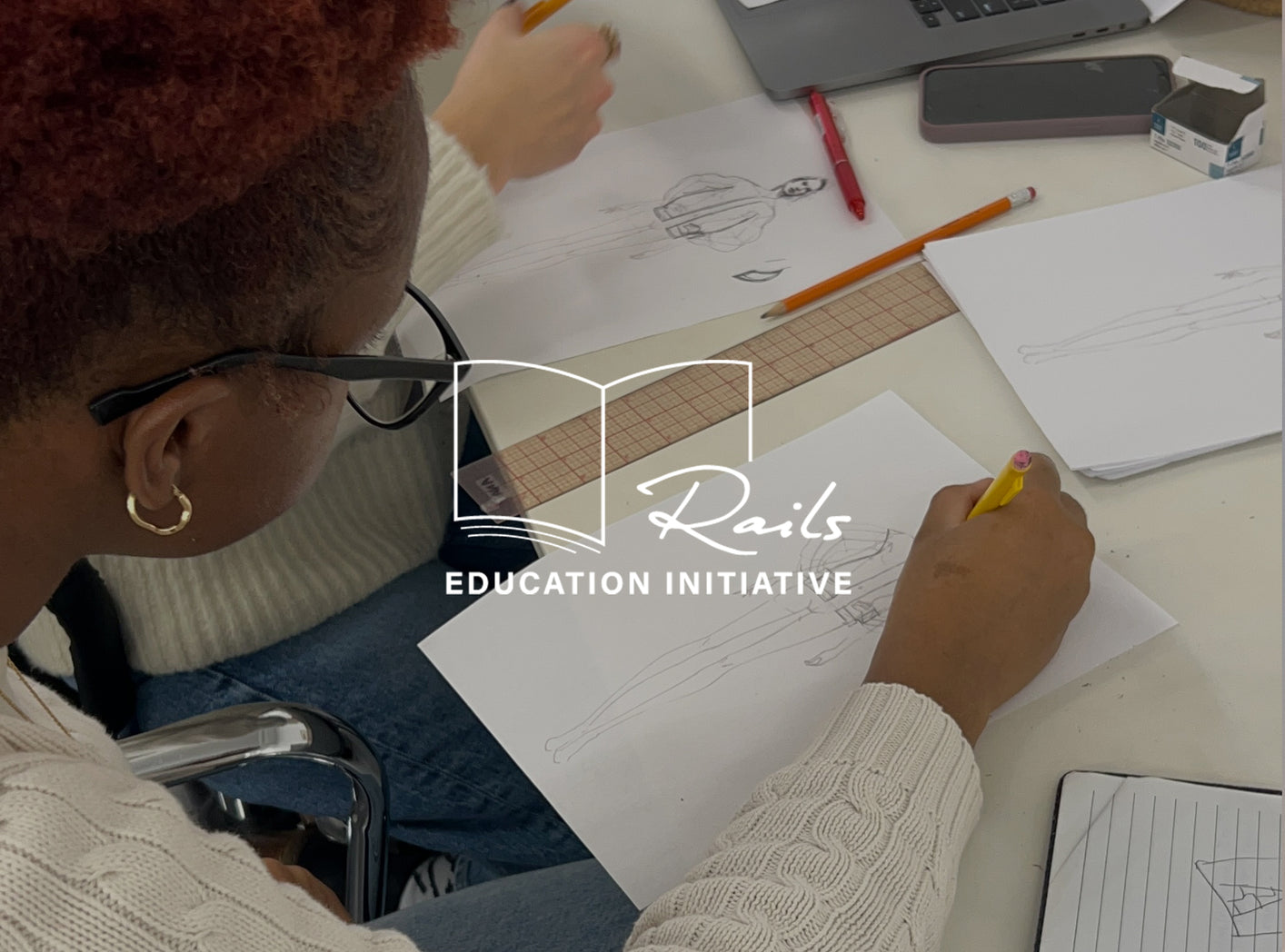 TWO FEMALES SITTING AT A DESK AND DRAWING SKETCHING OF CLOTHING ON WHITE PAPER