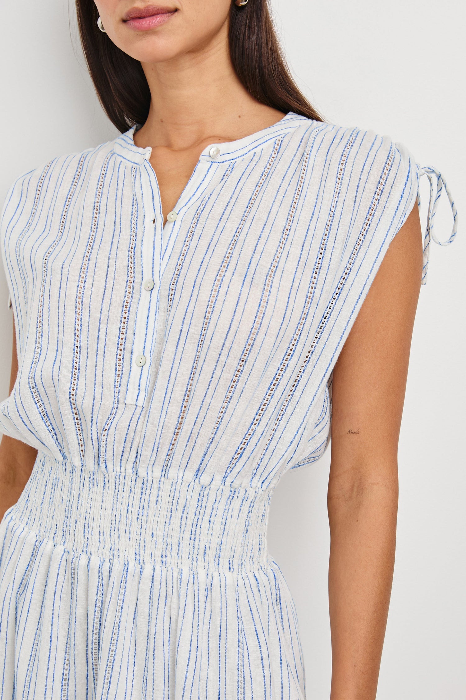 Samina Above-Knee Sleeveless Dress with drawstrings at the Sleeve Openings, Buttons along the Bodice, A Cinched Waist and a Tiered Skirt in Sonoma Stripe Colorway, Thin Vertical Blue Stripes on White - Front View Featured on Model 