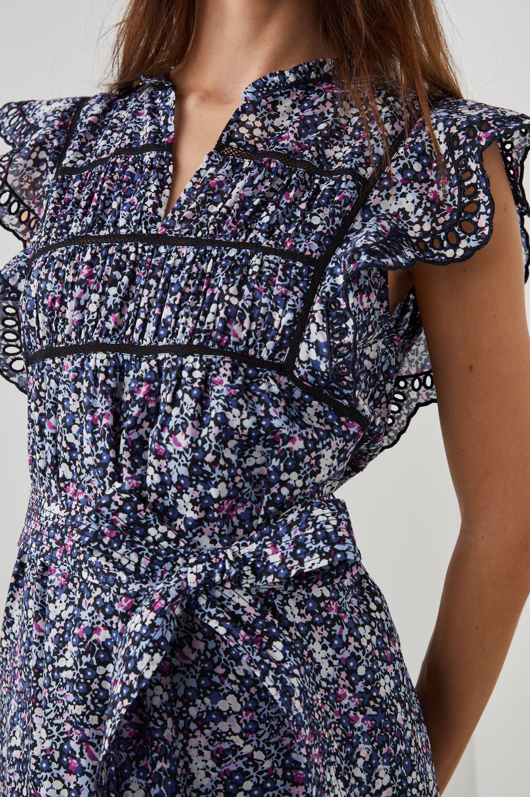 Sofie Short Sleeve Midi Dress with Flutter Sleeves, a V Neck, Panels with Shirring on the Bodice, a Fabric Waist Tie, a Tiered Skirt, and Cutouts on the Sleeves and Along the Hem in Midnight Hyacinth Colorway, Small Flowers in Blues, Pinks and White on Midnight - Front View Featured on Model 