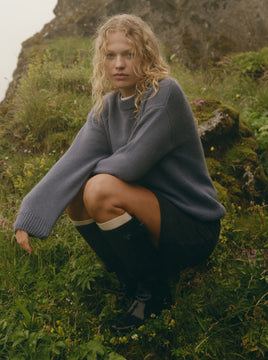 A woman crouching outdoors on a grassy hill wearing an oversized dusty blue-gray knit sweater, dark skirt, white socks, and black boots.