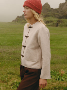 A person standing outdoors in a grassy field wearing a light beige wool-like jacket with dark toggle closures, dark brown trousers, and a red knit beanie.