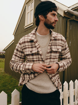 A man wearing a brown and beige plaid flannel shirt over a light gray knit sweater, with a black beanie and dark pants, standing outdoors in front of a rustic green wooden building and white picket fence.