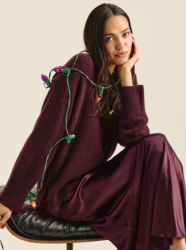 Woman seated on a black leather stool, chin resting on her hand, wearing a plum fuzzy knit sweater and a matching satin midi skirt, with a string of colorful vintage holiday lights draped over her.