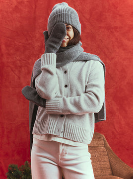 Smiling woman in a gray knit beanie, scarf, and mittens with a light gray button-up cardigan and white jeans, playfully covering one eye with her mitten against a red backdrop.