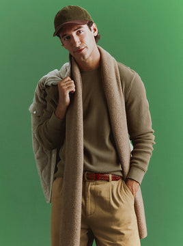 Man in an olive baseball cap and olive waffle-knit sweater with a tan knit scarf draped around his neck, a quilted jacket slung over his shoulder, khaki pants, and a woven leather belt, standing against a green backdrop.
