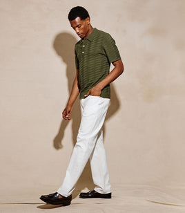 A man wearing a short-sleeve olive green striped polo shirt, white trousers, and black loafers walking against a warm beige studio backdrop.