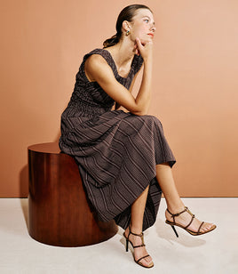 A woman sits on a dark wooden cylindrical stool against a warm peach studio backdrop, wearing a sleeveless dark midi dress with thin vertical pinstripes and strappy high-heeled sandals.