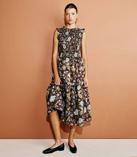 A woman models a sleeveless, high-neck midi dress with a brown floral print, standing against a solid terracotta-colored studio background.