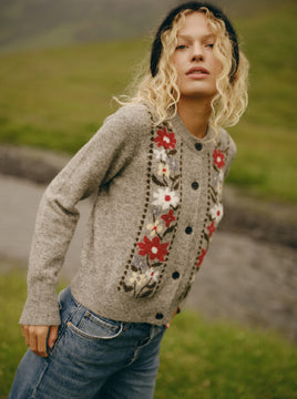 A woman wearing a heather gray button-up cardigan with a vertical floral pattern in red, white, and black, paired with blue jeans and a black beanie, set against a blurred green outdoor background.