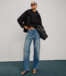 Person wearing a black outfit with blue jeans and sunglasses, holding a black bag against a white background.