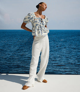 Woman in a floral top and white pants standing on a dock with ocean in the background