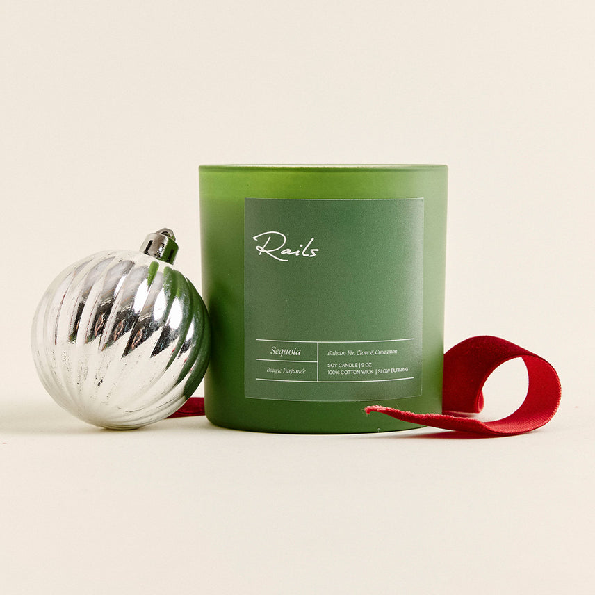 Green frosted Rails Sequoia soy candle with balsam fir, clove, and cinnamon scent, beside a silver ribbed ornament and a red velvet ribbon.